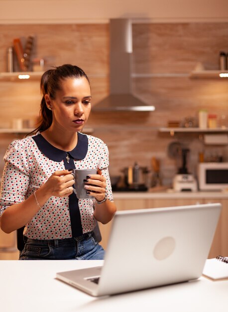 Donna seduta in cucina lat di notte lavorando su un progetto di lavoro utilizzando laptop e tenendo una tazza di caffè. Dipendente che utilizza la tecnologia moderna a mezzanotte facendo gli straordinari per lavoro, affari, impegni, carriera.