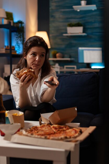 Donna seduta sul divano che mangia gustoso hamburger delizioso con telecomando che cambia canale in televisione davanti al tavolo con pizza grande. Persona che naviga nelle stazioni TV mentre mangia hamburger.