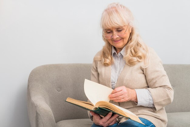 Donna senior allegra che si siede sul sofà che legge il libro