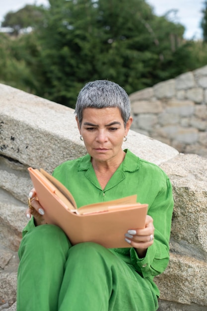 Donna senior assorta che legge nel parco. Modello femminile con capelli grigi corti in abiti luminosi che tengono libro. Tempo libero, concetto di letteratura