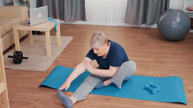 Donna senior attiva che allunga corpo sulla stuoia di yoga. Il pensionato anziano esercita l'allenamento a casa attività sportiva all'età pensionabile anziana elderly