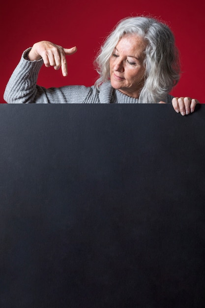 Donna senior che indica il suo dito verso il basso sul cartello nero in bianco contro il contesto rosso