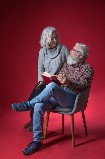 Donna senior che si siede con il suo marito che si siede sulla sedia che giudica il libro a disposizione contro fondo rosso