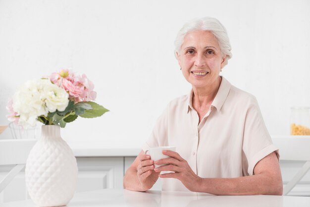 Donna senior che tiene tazza di caffè con il vaso di fiore sulla tavola bianca