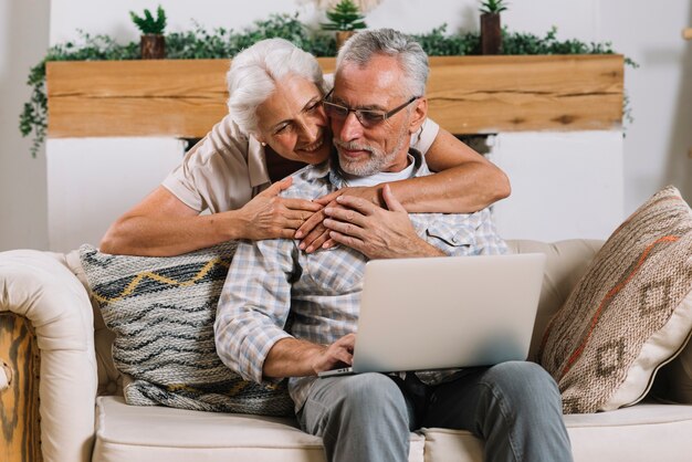 Donna senior felice che abbraccia il suo marito da dietro che si siede sul sofà con il computer portatile