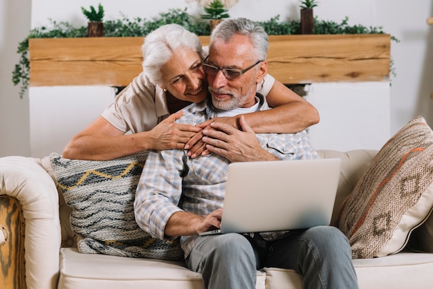 Donna senior felice che abbraccia il suo marito da dietro che si siede sul sofà con il computer portatile