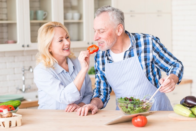 Donna senior felice che alimenta la fetta rossa del peperone dolce al suo marito che prepara l&#39;insalata nella cucina