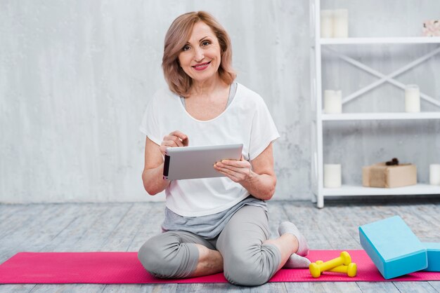 Donna senior felice che per mezzo del computer portatile vicino all&#39;attrezzatura di yoga