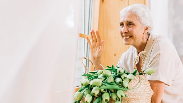 Donna senior sorridente che osserva attraverso la finestra che fluttua la sua mano