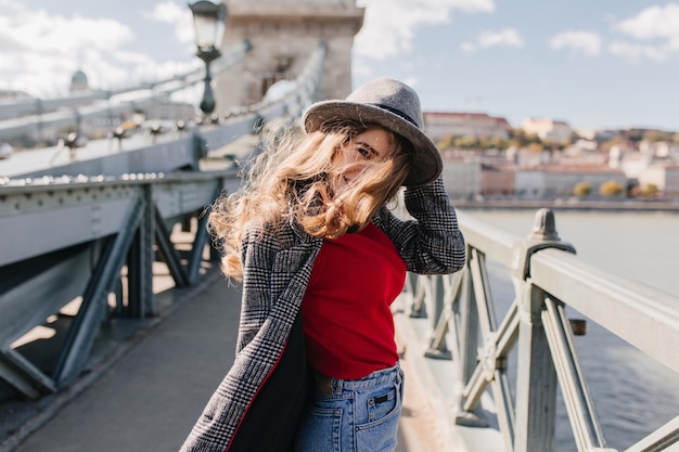 Donna sensuale castana dai capelli lunghi che sorride giocosamente durante il viaggio in città europea. Ritratto all'aperto di una ragazza straordinaria in abiti a maglia rossa che esprime felicità mentre si trova sul ponte.