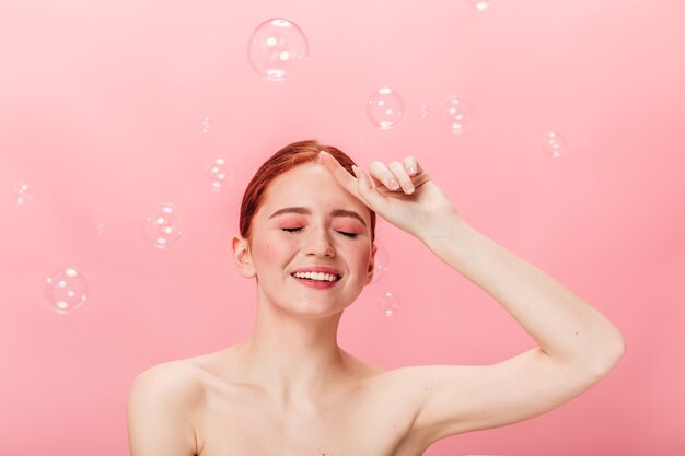 Donna sensuale dello zenzero che posa con le bolle di sapone. Eccitato ragazza nuda ridendo isolato su sfondo rosa.