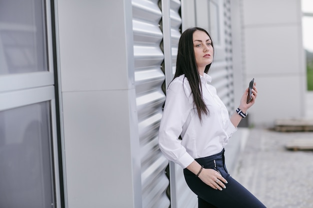 Donna seria appoggia su una parete bianca con un telefono in mano