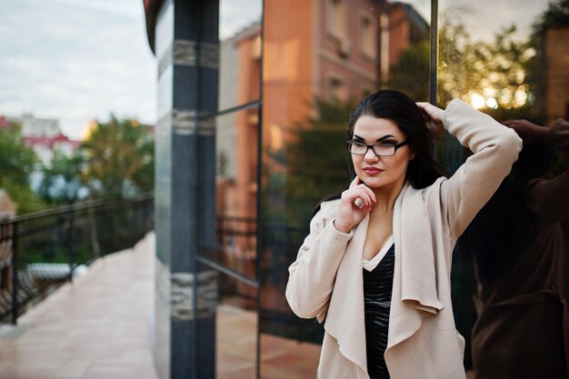 Donna sexy capelli neri in occhiali e cappotto poste contro edificio con finestre moderne