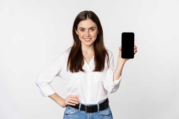 Donna sicura di sé in abiti aziendali, che mostra lo schermo dello smartphone, l'interfaccia mobile di un'applicazione, in piedi su sfondo bianco.