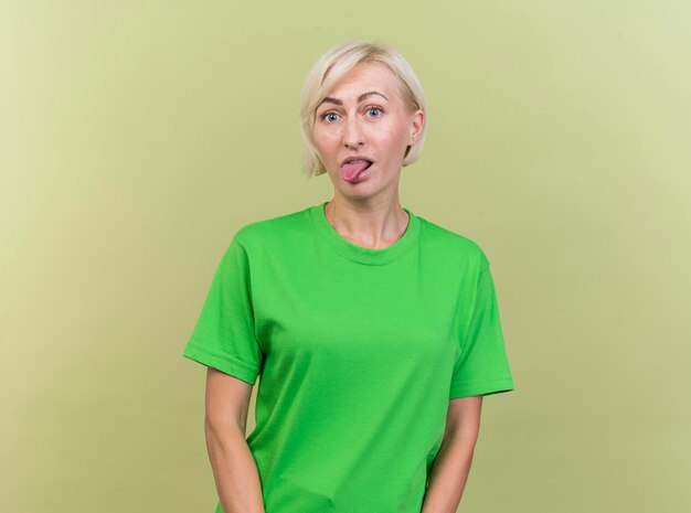 Donna slava bionda di mezza età allegra che esamina la linguetta che mostra anteriore isolata sulla parete verde oliva con lo spazio della copia