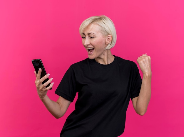 Donna slava bionda di mezza età allegra che tiene guardando il telefono cellulare che fa gesto di sì isolato sulla parete rosa