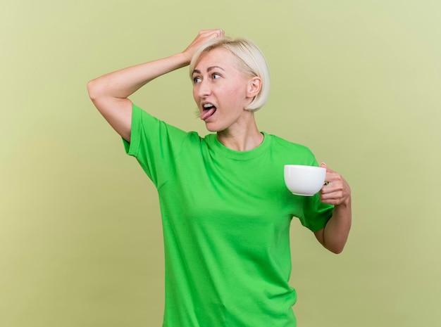 Donna slava bionda di mezza età irritata che tiene la tazza di tè che tocca la testa che guarda il lato che mostra la linguetta isolata sulla parete verde oliva