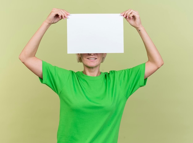 Donna slava bionda di mezza età sorridente che tiene documento in bianco davanti agli occhi isolati sulla parete verde oliva