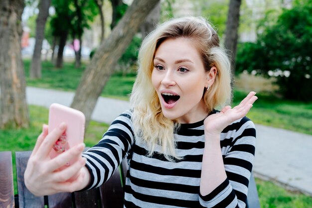 Donna sorpresa che prende selfie con lo smartphone mentre sedendosi all'aperto su un banco nella cima a strisce