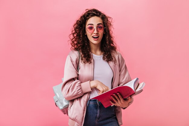 Donna sorpresa in giacca rosa e jeans in posa con libri su sfondo isolato