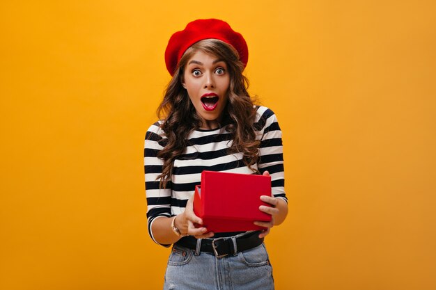 Donna sorpresa in scatola regalo apertura berretto rosso su sfondo arancione. Bella ragazza con acconciatura ondulata in cappello luminoso e abiti moderni esulta.