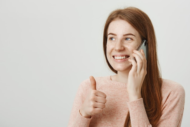 Donna sorridente attactive della testarossa che parla sul telefono cellulare e mostra il pollice in su