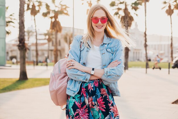 donna sorridente che cammina in una strada cittadina in elegante gonna stampata e giacca oversize in denim indossando occhiali da sole rosa