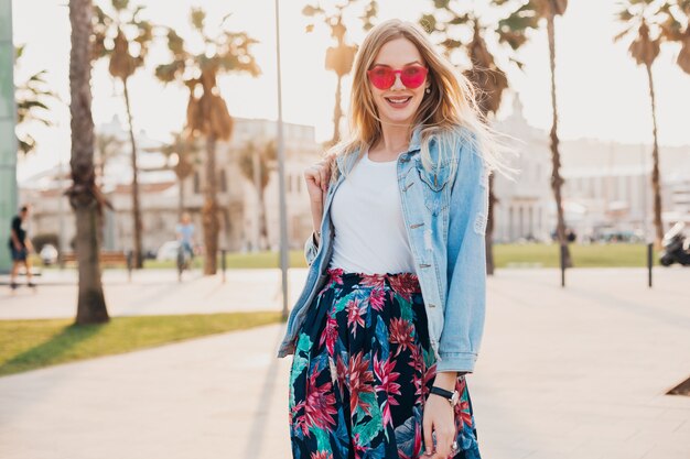 donna sorridente che cammina in una strada cittadina in elegante gonna stampata e giacca oversize in denim indossando occhiali da sole rosa