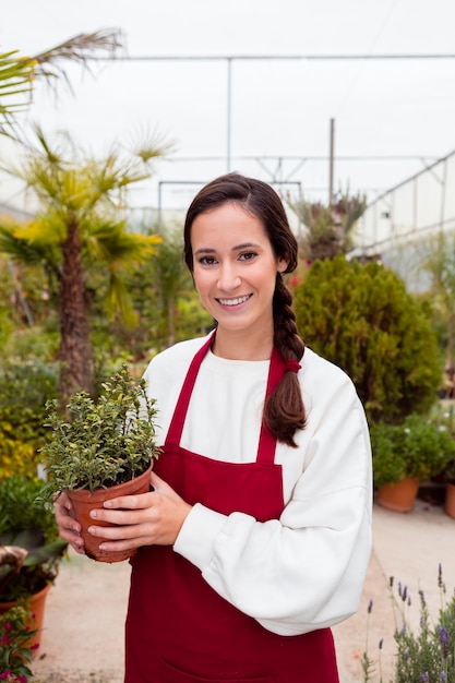 Donna sorridente che indossa i vestiti di giardinaggio e che tiene vaso in serra