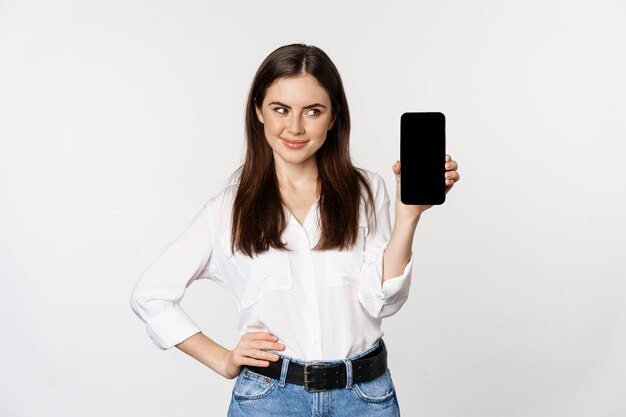Donna sorridente che mostra lo schermo del telefono cellulare, cercando astuta, in piedi su sfondo bianco