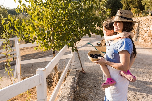 Donna sorridente che porta sua figlia che tiene le olive raccolte nella ciotola