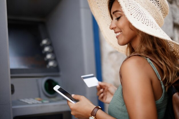 Donna sorridente che preleva denaro dal bancomat durante l'utilizzo di smartphone
