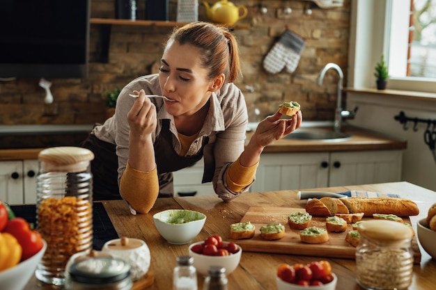 Donna sorridente che si gode il gusto del cibo sano mentre prepara la bruschetta all'avocado in cucina