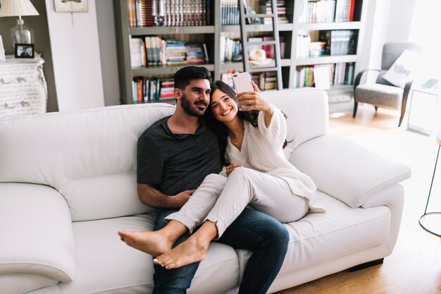 Donna sorridente che si siede con il suo ragazzo che prende selfie sul cellulare a casa