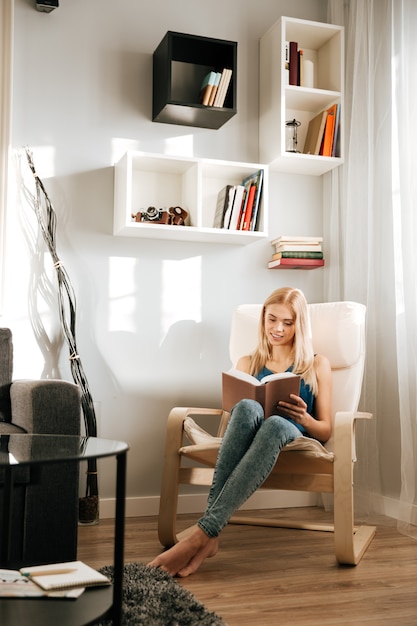 Donna sorridente che si siede in sedia e libro di lettura a casa