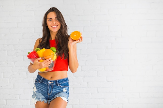 Donna sorridente che sta contro la parete che tiene frutta e verdura fresca