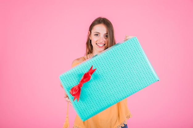 Donna sorridente che tiene il grande contenitore di regalo con l&#39;arco rosso contro fondo rosa