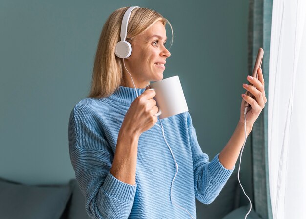 Donna sorridente che utilizza smartphone e cuffie a casa mentre beve il caffè durante la pandemia