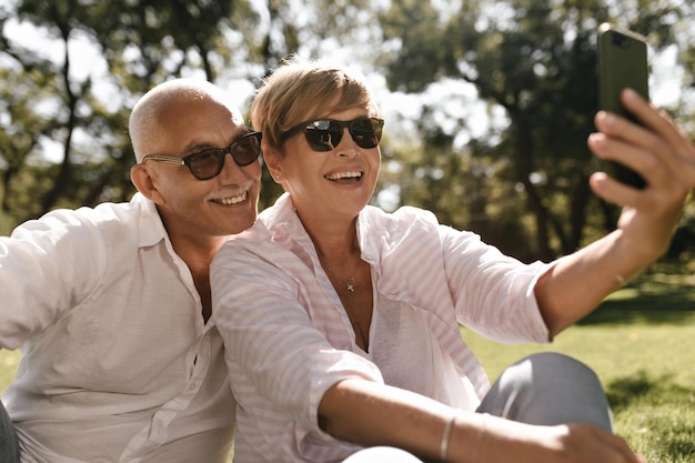 Donna sorridente con l'acconciatura moderna bionda in abiti leggeri a strisce che fanno selfie con l'uomo in occhiali da sole e camicia bianca all'aperto