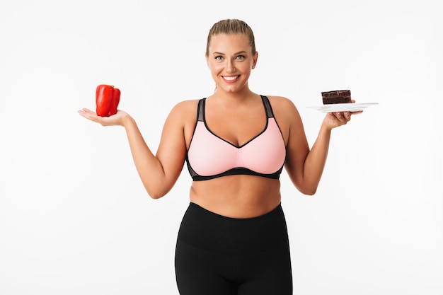 Donna sorridente con peso in eccesso nella parte superiore sportiva che tiene in mano pepe rosso e torta al cioccolato mentre guarda felicemente a porte chiuse su sfondo bianco