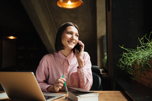 Donna sorridente del brunette che si siede dalla tabella con il computer portatile