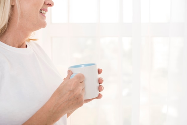 Donna sorridente del primo piano con la tazza vicino alla finestra