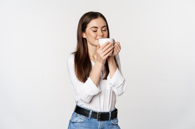 Donna sorridente dell'ufficio che beve caffè, beve dalla tazza con soddisfazione, piacere, in piedi su sfondo bianco