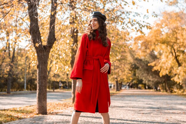 Donna sorridente elegante attraente con capelli ricci che cammina nel parco vestita di moda alla moda autunno caldo cappotto rosso, street style, indossando il cappello berretto