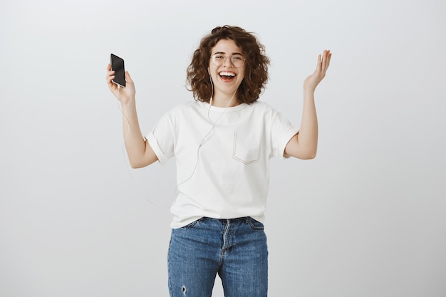 Donna sorridente felice che alza le mani in rallegrarsi mentre ascolta musica in cuffia e tiene smartphone