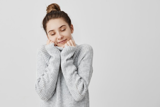 Donna sorridente felice che avvolge il suo viso nel colletto del maglione grigio. Cutie femminile sentirsi a proprio agio e calda prendendo piacere chiudendo gli occhi. Concetto di armonia