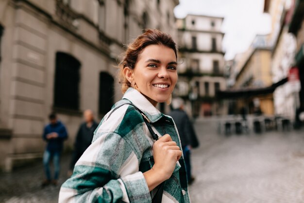 Donna sorridente felice con capelli biondi che indossa la camicia a righe che cammina sulla vecchia strada soleggiata. La ragazza con la borsa sta camminando in città