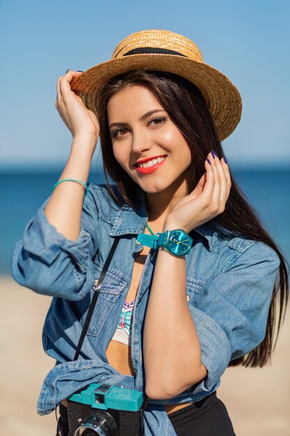 Donna sorridente in cappello di paglia e vestito estivo alla moda in posa con la retro macchina fotografica sulla spiaggia.