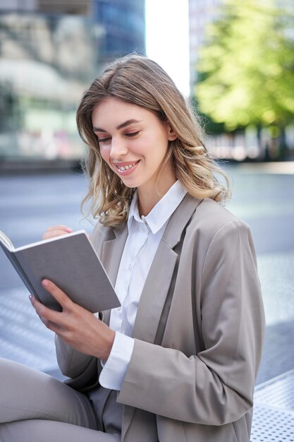 Donna sorridente in giacca e cravatta che scrive idee usando taccuino e penna seduti all'aperto nel lavoro del centro città