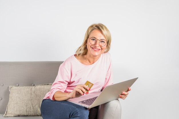 Donna sorridente invecchiata in camicetta rosa con carta di plastica e laptop sul divano